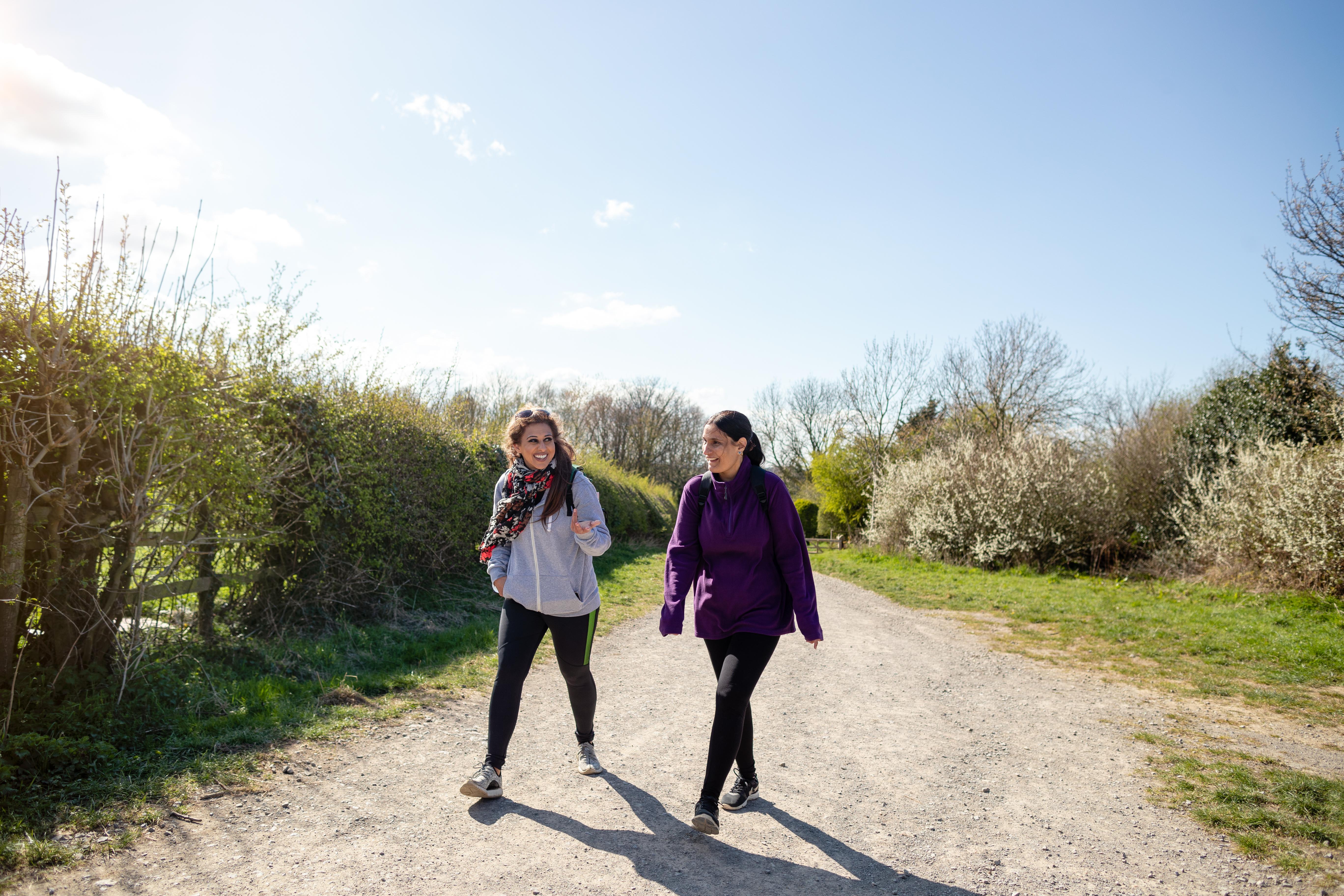 Two women walk side by side along a gravel road, sunshine, blue skies and green hedges on either side