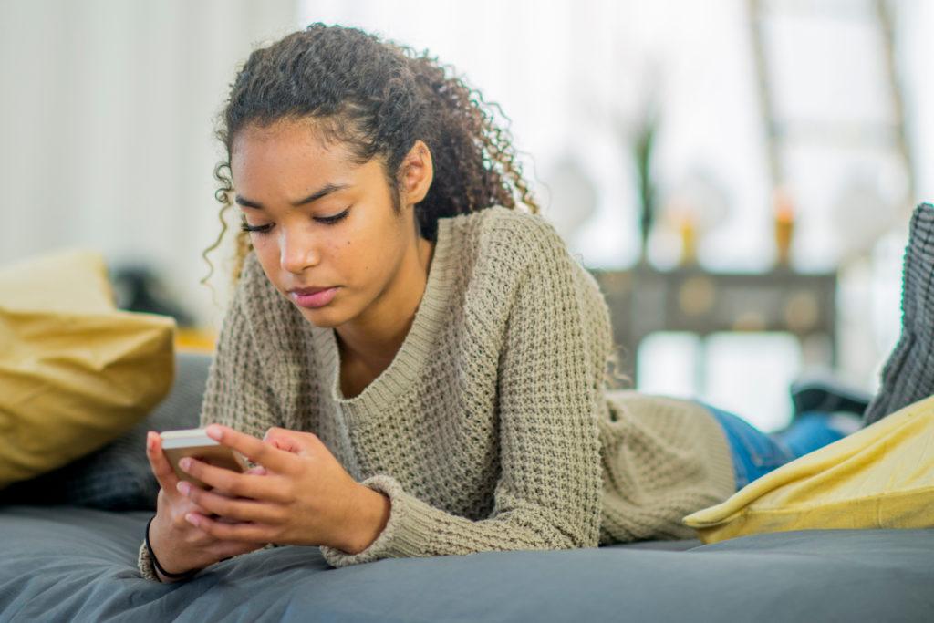 A teenage girl texts on her phone while lying on her bed.