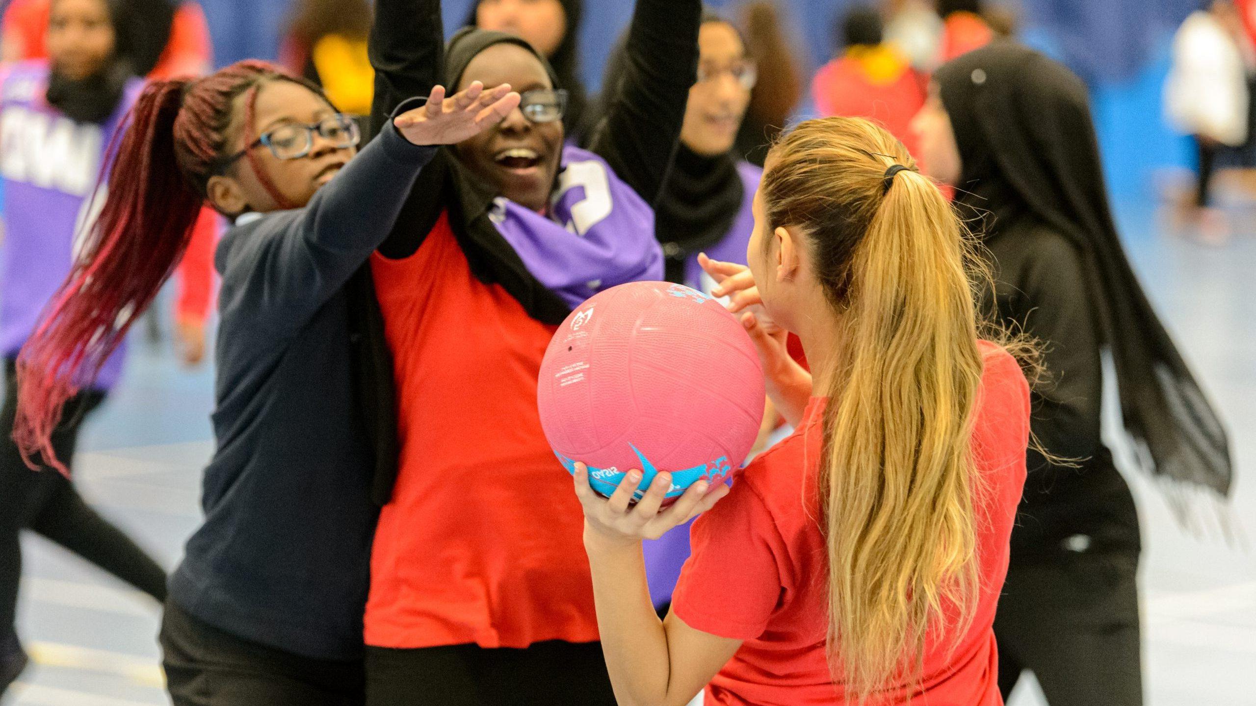 A group of high school girls playing netball. A white girl is passing the ball to a black girl in her team