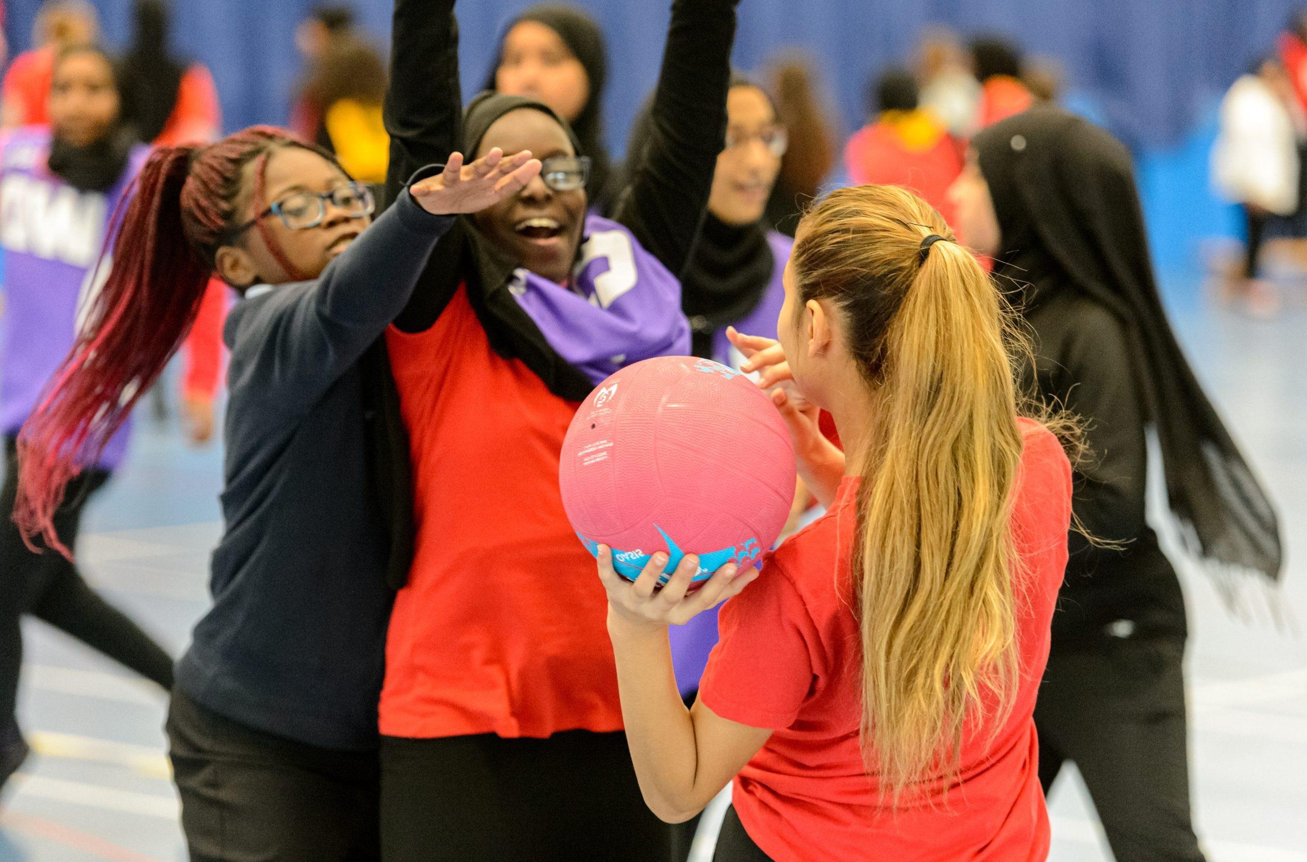 A group of high school girls playing netball. A white girl is passing the ball to a black girl in her team