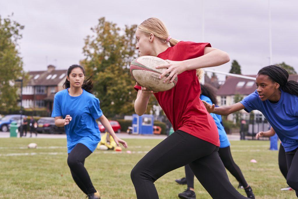 White teenage girl wearing a read t shirt and black leggings running with a rugby ball 