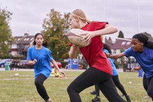 White teenage girl wearing a red t shirt and black leggings running with a rugby ball